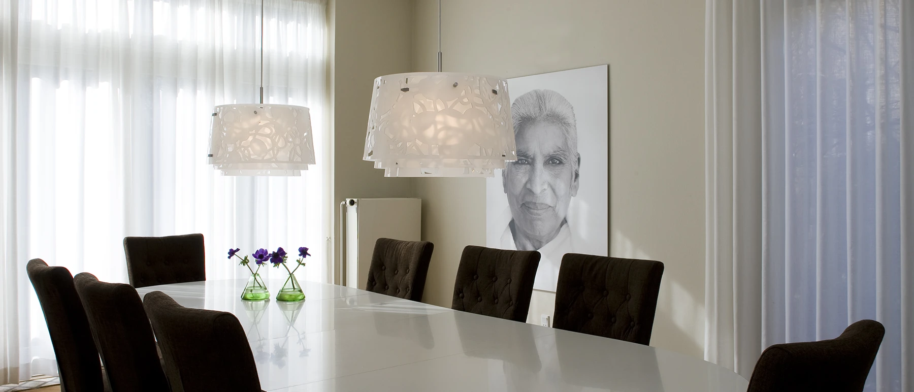 Two white Collage Pendants hang over a dining table with dark chairs in a bright room. © 2026 Louis Poulsen.