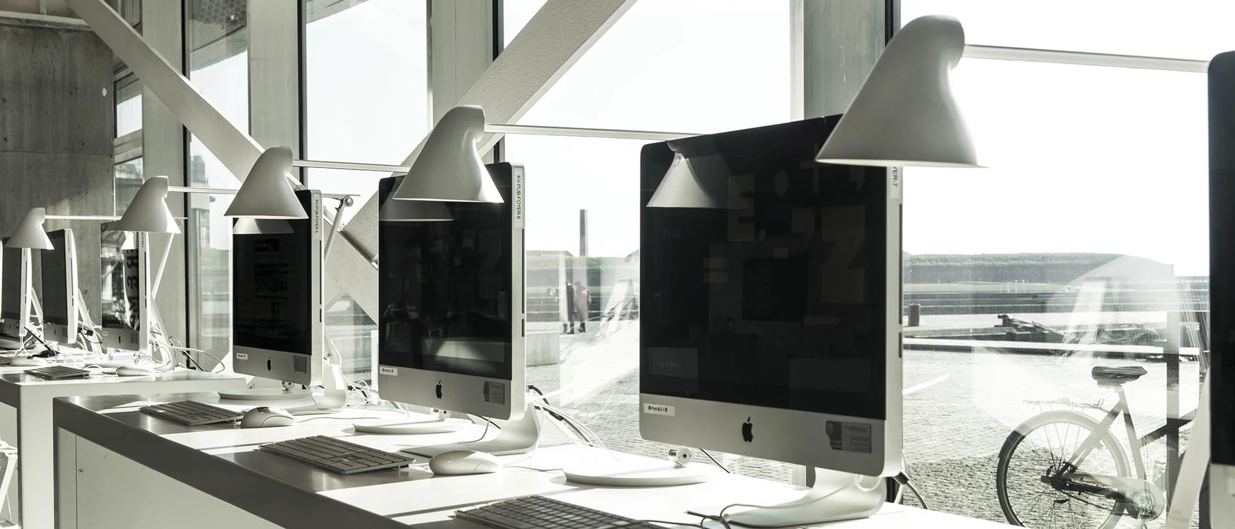 White NJP Table Lamps sit beside computer screens in a bright office with large windows. © 2026 Louis Poulsen.