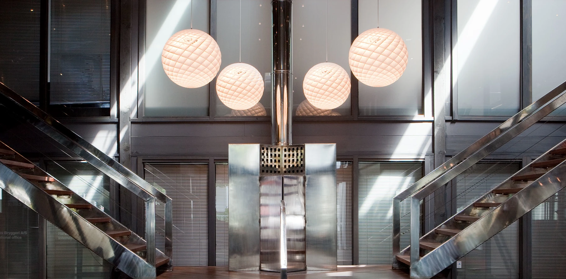Four glowing Patera pendants hang in a modern atrium with metal staircases and large windows. © 2026 Louis Poulsen.