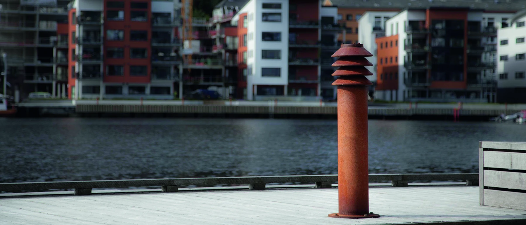 Eine rostfarbene Bysted Pollerleuchte steht auf einer Holzpromenade am Wasser, dahinter moderne Wohnhäuser. © 2026 Louis Poulsen.