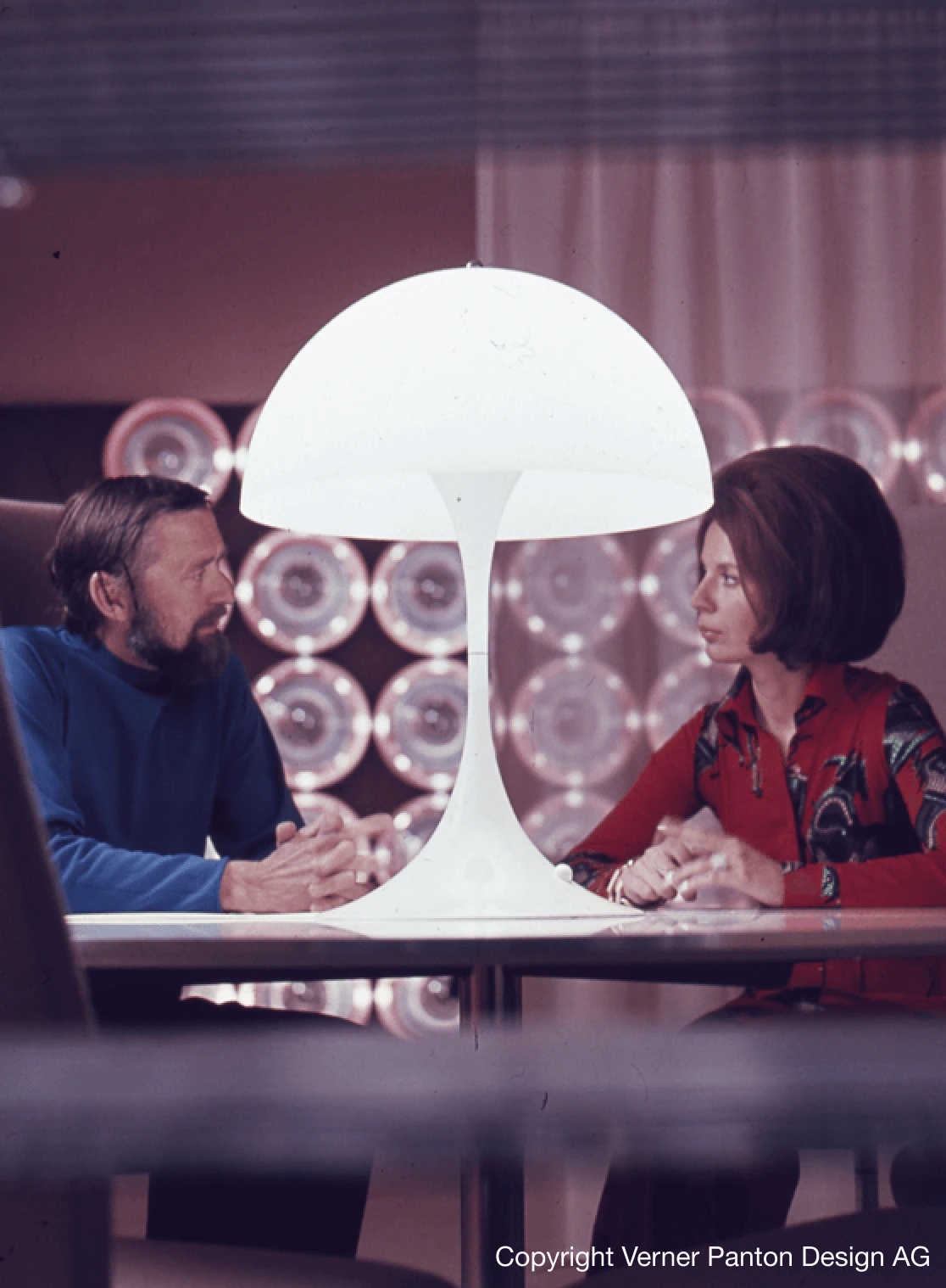 Una Panthella lámpara de pie blanca está sobre una mesa entre dos personas, con luces circulares al fondo. © 2026 Louis Poulsen.