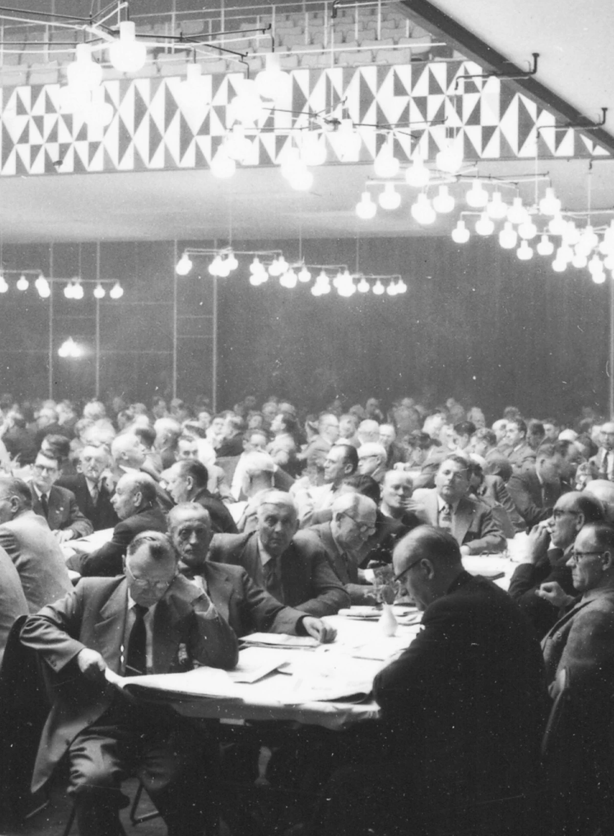 VL 45 Radiohus-pendler lyser opp en fullsatt møtesal med lange bord og geometrisk veggdekor. © 2026 Louis Poulsen.