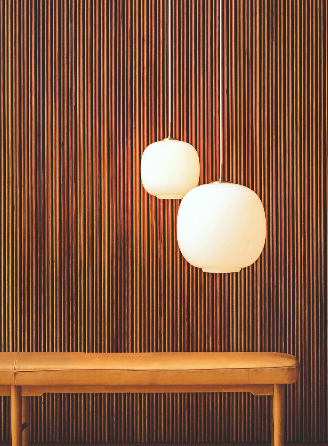 To VL 45 Radiohus-pendler i opalglass lyser foran en mørk vegg med trespiler, over en benk i skinn. © 2026 Louis Poulsen.