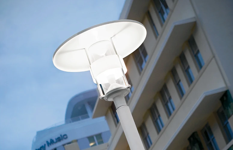 Une borne Albertslund Maxi Post éclaire les abords d’un immeuble moderne sous un ciel bleu. © 2026 Louis Poulsen.