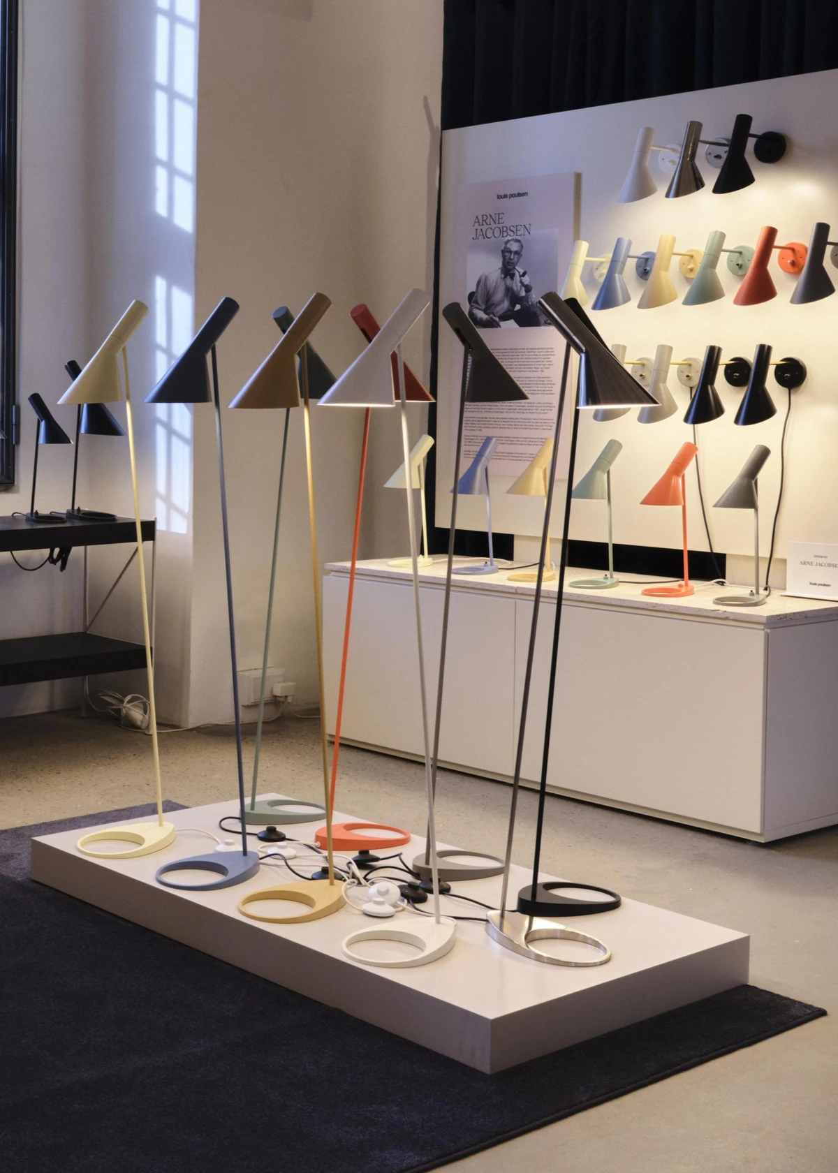 Kleurrijke AJ Floor Lamp vloerlampen staan op een podium in de showroom in Kopenhagen, met AJ Wall Lamp wandlampen aan de muur. © 2026 Louis Poulsen.