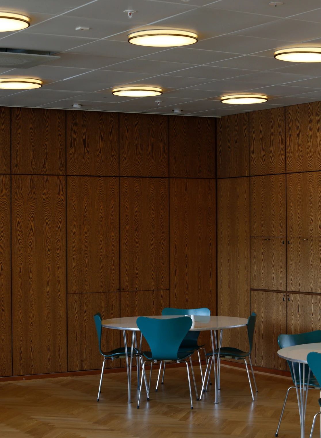 AJ Munkegaard plafondlampen verlichten een vergaderruimte met houten wandpanelen, parketvloer en blauwe stoelen. © 2026 Louis Poulsen.