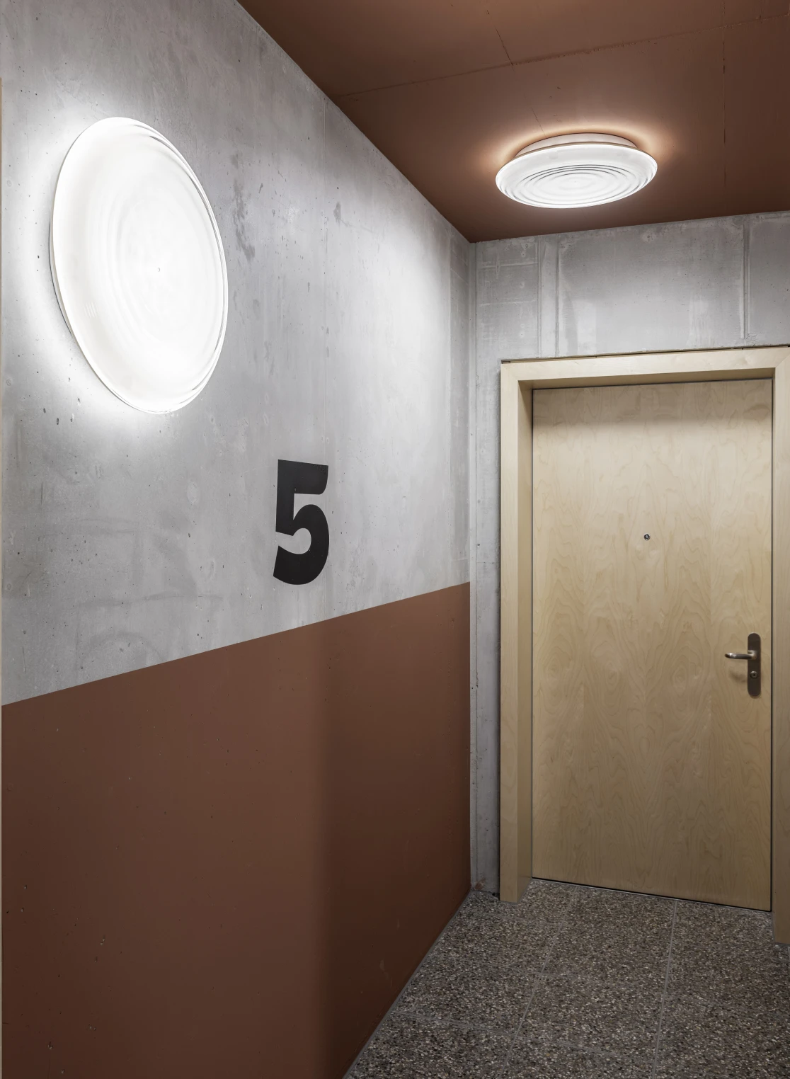 Ripls wand- en plafondlampen verlichten een betonnen gang naast een houten deur met het nummer 5. © 2026 Louis Poulsen.