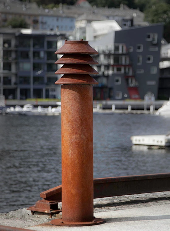 A rust-coloured Bysted Bollard stands beside the water, with modern buildings in the background. © 2026 Louis Poulsen.