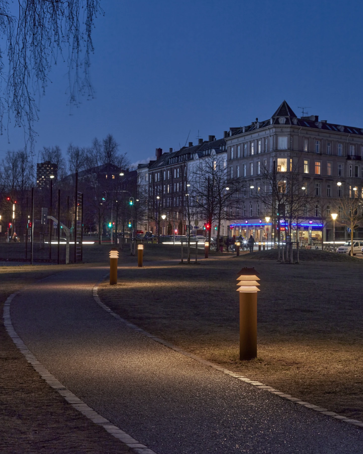 Ballerup-bolderarmaturen verlichten een slingerend pad in een stadspark, met historische gebouwen op de achtergrond. © 2026 Louis Poulsen.
