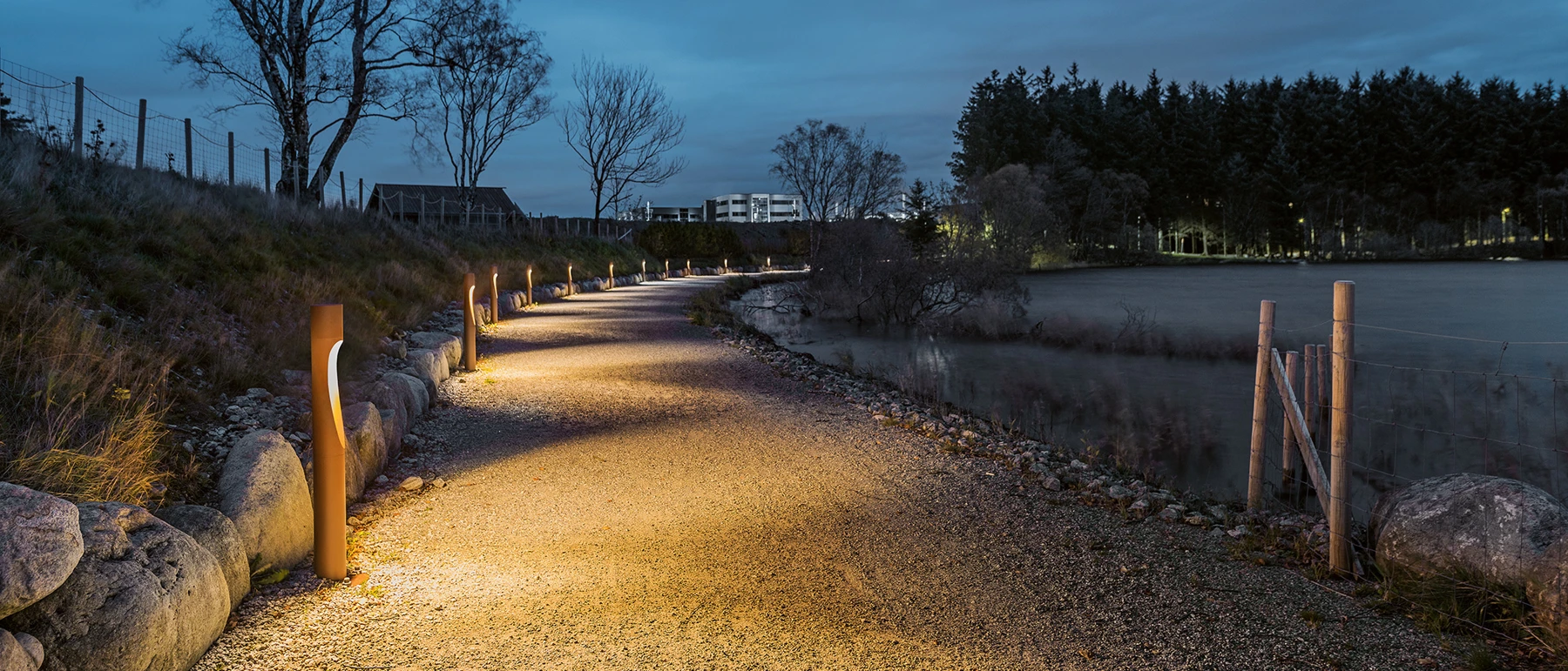 Flindt Bollards in corten steel illuminate a gravel path beside the lake at dusk. © 2026 Louis Poulsen.