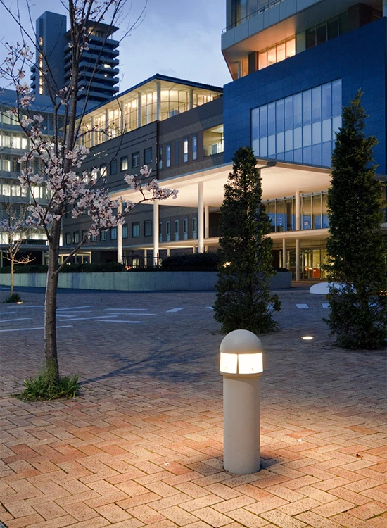 A white Albertslund Mini Post lights the paved forecourt of a modern hospital at dusk. © 2026 Louis Poulsen.