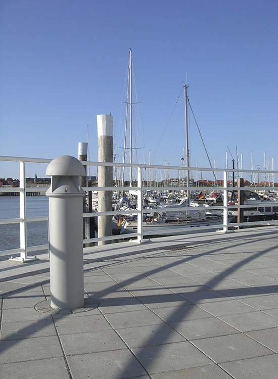 A grey Albertslund Maxi Post on a harbour promenade, with sailboats, mooring posts and a clear sky. © 2026 Louis Poulsen.