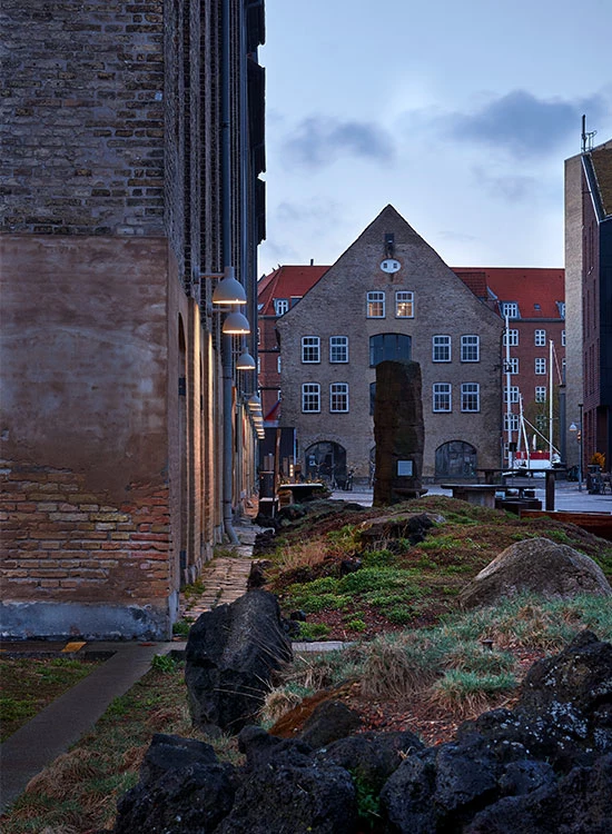 Toldbod 155 buitenwandlampen verlichten een bakstenen gevel bij een groen ingerichte stadsruimte. © 2026 Louis Poulsen.