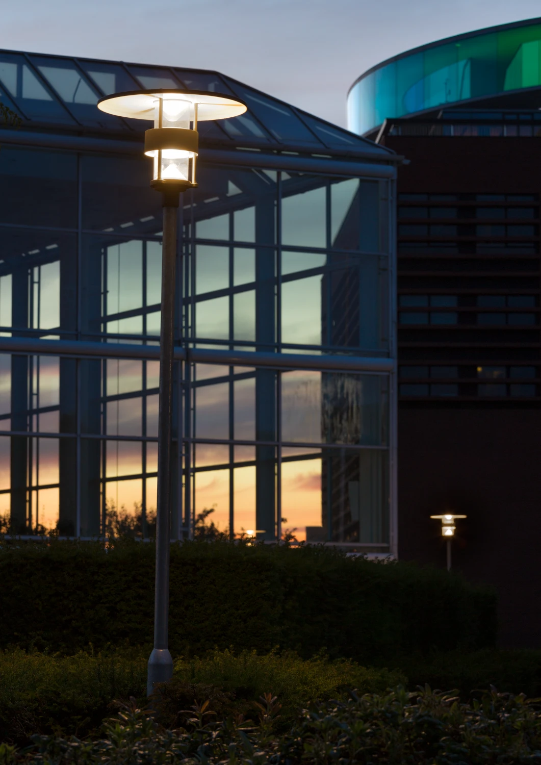 Albertslund Maxi Post-lantaarns verlichten bij zonsondergang de buitenruimte voor de glazen gevel van Musikhuset Aarhus. © 2026 Louis Poulsen.