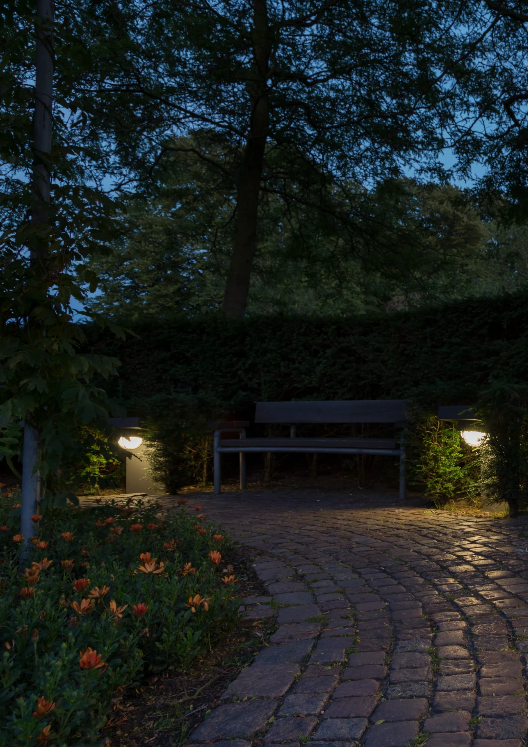 Flindt Bollard verlicht in de schemering een geplaveid tuinpad met bloemen, een haag en een bankje. © 2026 Louis Poulsen.