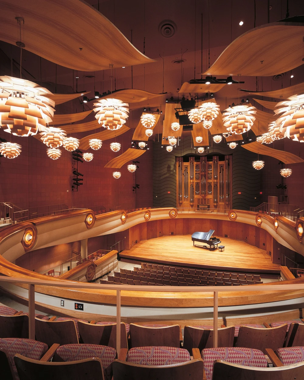 PH Artichoke hanglampen verlichten het podium van een concertzaal met gebogen houten panelen en rijen stoelen. © 2026 Louis Poulsen.