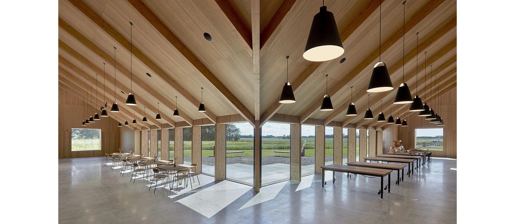 Black Above Pendants hang over tables in Kornets Hus, a bright hall with a timber ceiling and views of fields. © 2026 Louis Poulsen.