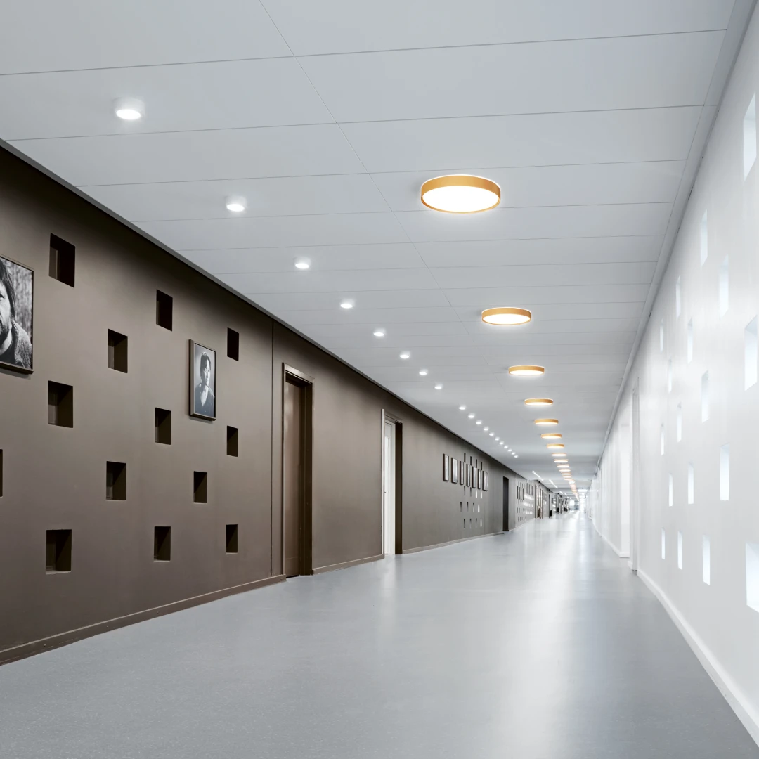 LP Circle Surface Mounted plafondarmaturen verlichten een lange, moderne ziekenhuisgang met lichte muren en een grijze vloer. © 2026 Louis Poulsen.