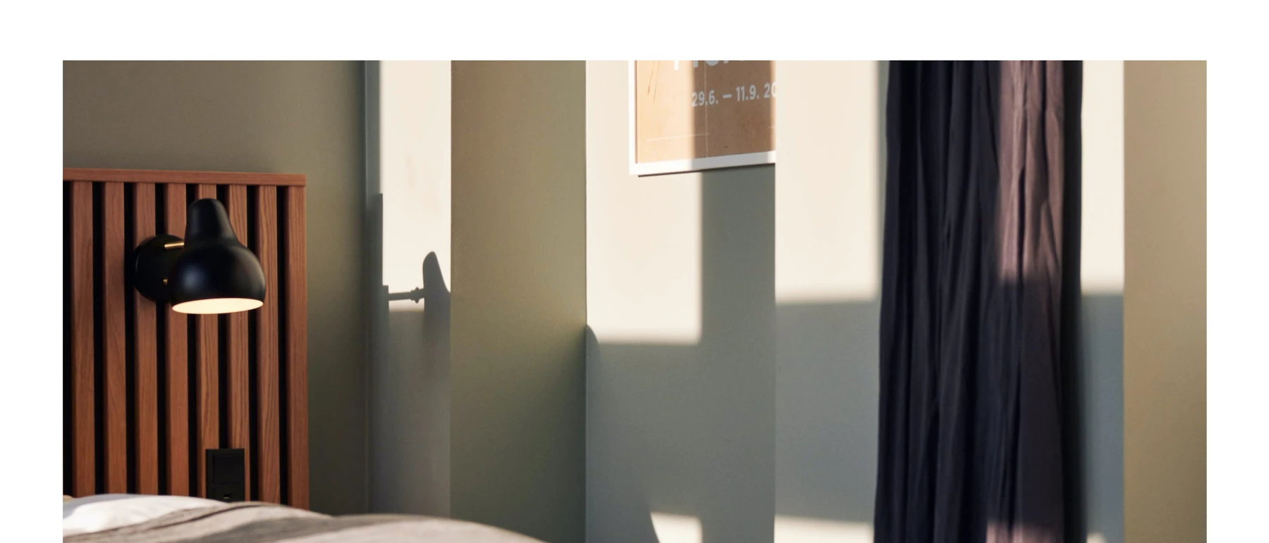 A black AJ Wall Lamp beside a bed with a slatted wooden headboard at Coco Hotel, lit by sunlight. © 2026 Louis Poulsen.