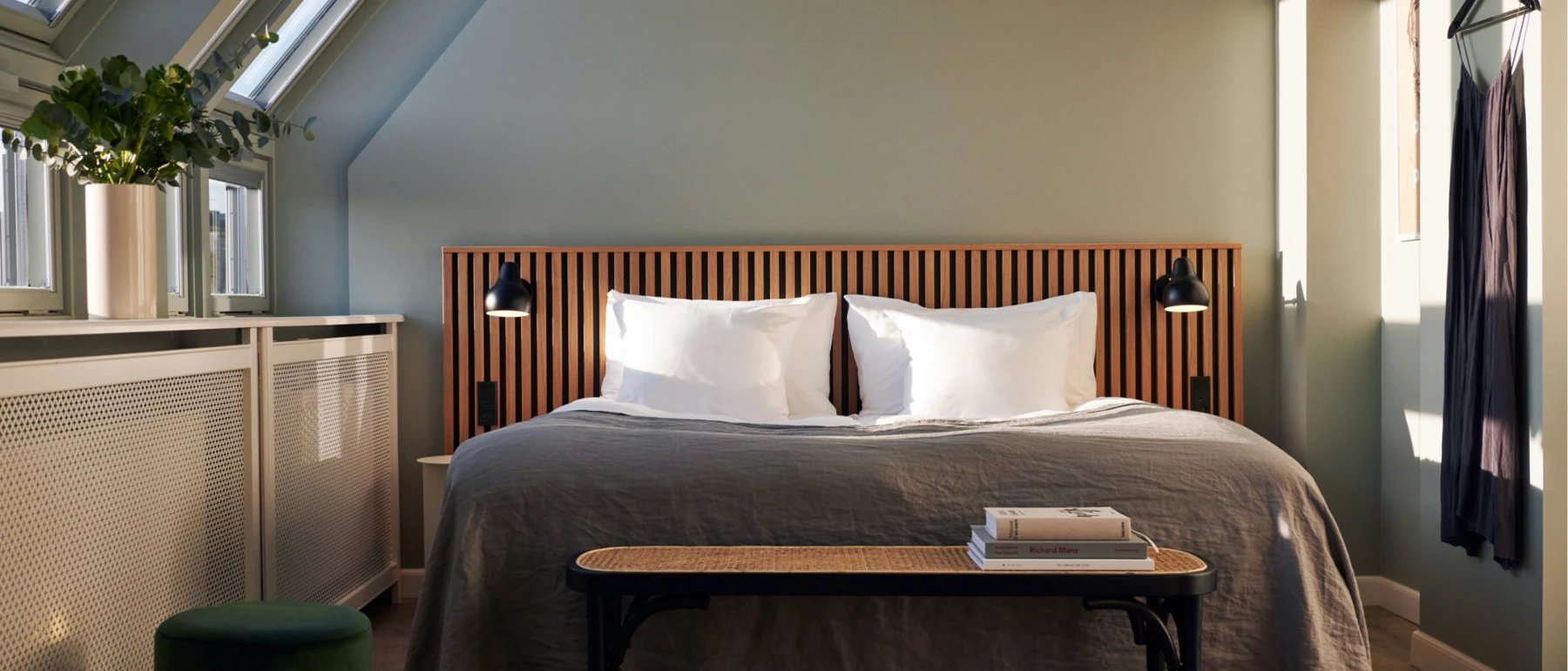 Two black AJ Wall Lamp wall lights flank a bed in a bright hotel room with a slatted wooden headboard. © 2026 Louis Poulsen.