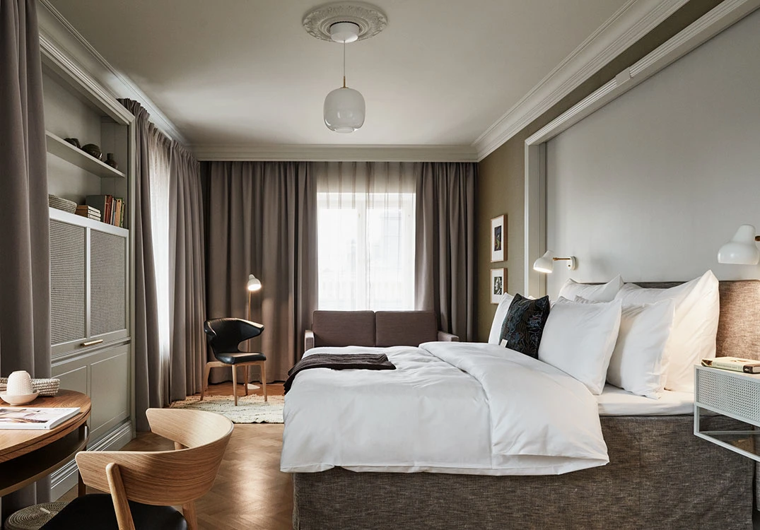 A white Wohlert Pendant and AJ Wall Lamp illuminate an elegant hotel room in muted tones. © 2026 Louis Poulsen.