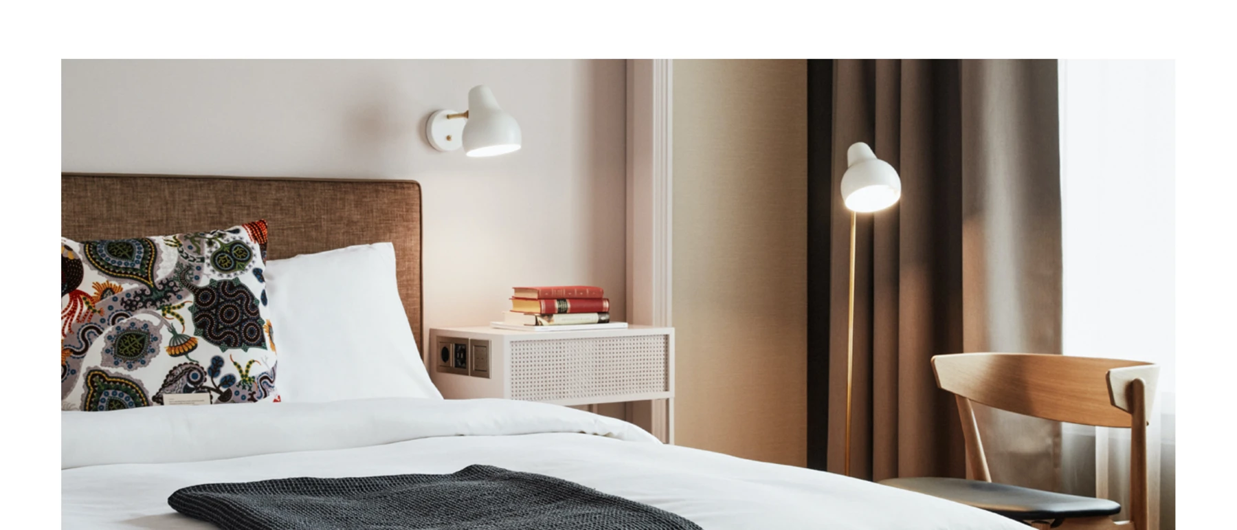 A white AJ Wall Lamp and AJ Floor Lamp illuminate a hotel bed in a room with warm, neutral tones. © 2026 Louis Poulsen.