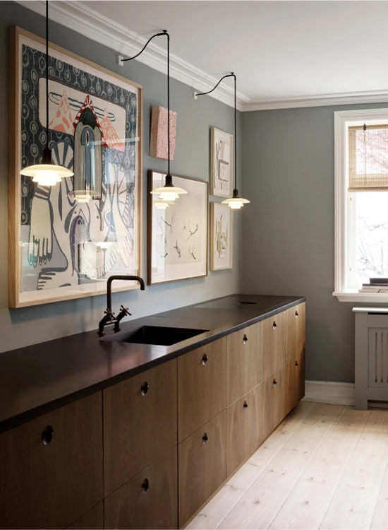 Three PH 2/1 Pendant lights with white shades hang above a dark countertop in a kitchen with wooden cabinets. © 2026 Louis Poulsen.