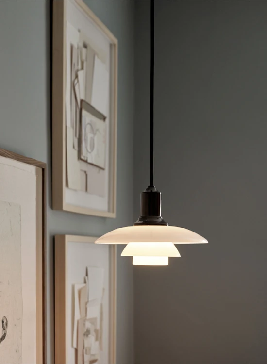 A PH 3½-3 Pendant with white shades casts a warm glow beside framed art on a dark grey wall. © 2026 Louis Poulsen.