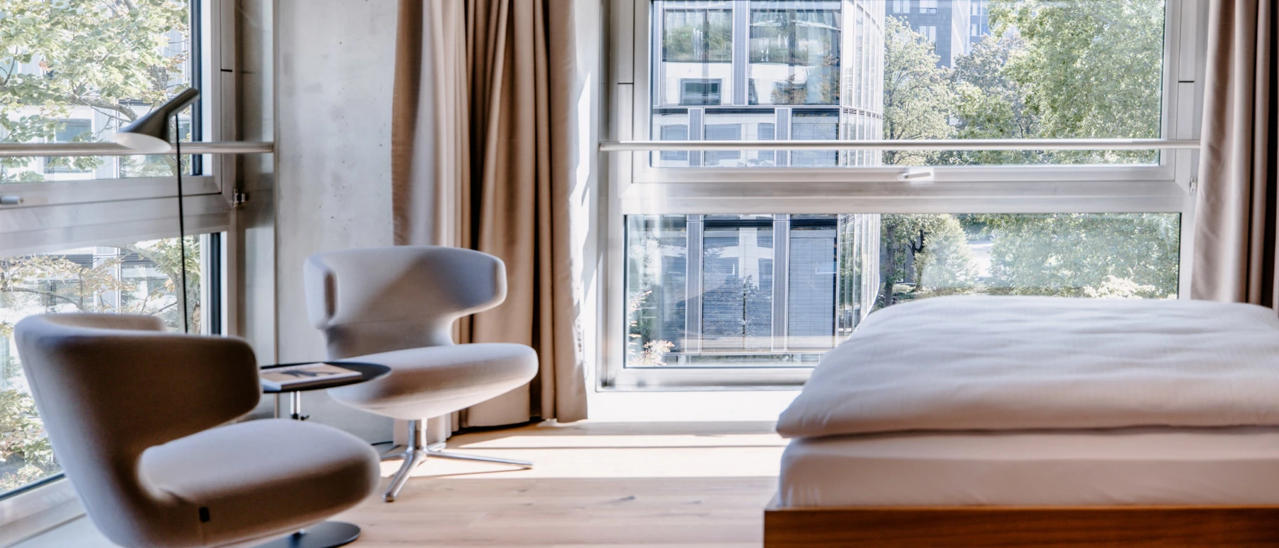 A black AJ Floor Lamp stands beside lounge chairs in a bright hotel room with large windows overlooking the city. © 2026 Louis Poulsen.