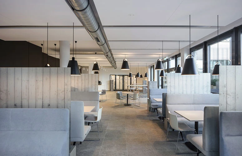 Black Toldbod Pendants hang above seating areas in a bright office with large windows. © 2026 Louis Poulsen.