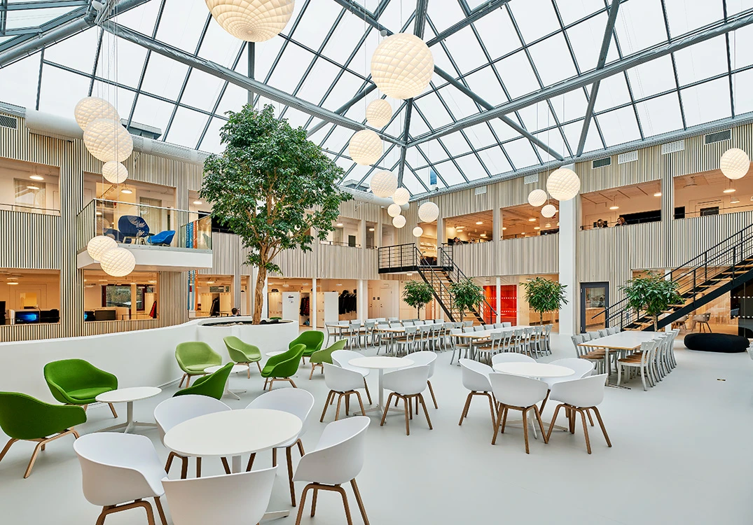 Patera-Pendelleuchten erhellen das helle Büroatrium von Kraftringen mit Glasdach, grünen Stühlen und Gemeinschaftstischen in Lund. © 2026 Louis Poulsen.