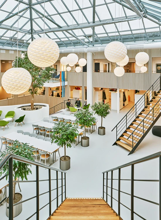 Weiße Patera Pendelleuchten hängen im hellen Atrium des Kraftringen-Büros mit Glasdach, Pflanzen und Treppen. © 2026 Louis Poulsen.