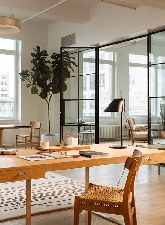 An AJ Table Lamp in a copper finish sits on a desk in a bright office with wooden furniture. © 2026 Louis Poulsen.