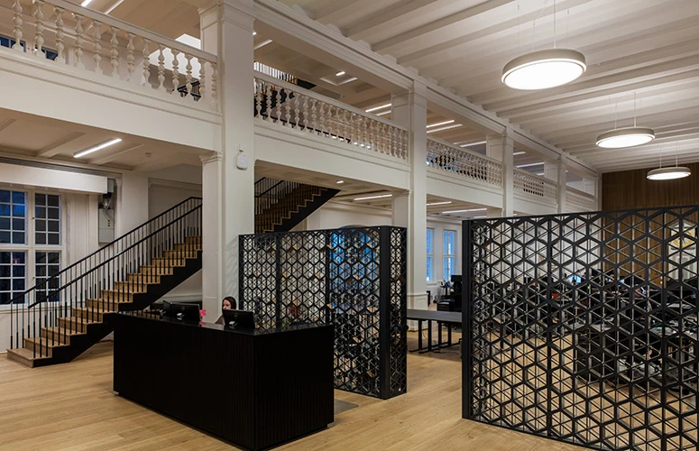 LP Grand Suspended pendants light Momondo’s office, with pale wood floors, a staircase and black lattice dividers. © 2026 Louis Poulsen.