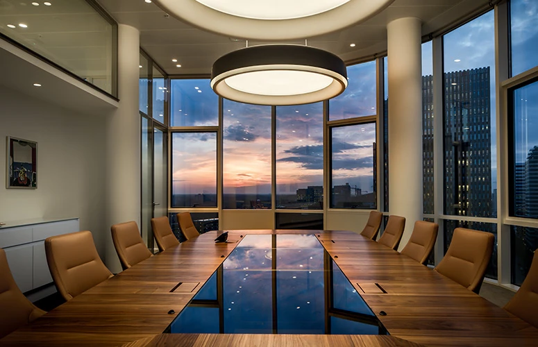 Round LP Grand ceiling lamps illuminate a conference room with long wooden tables and city views at sunset. © 2026 Louis Poulsen.
