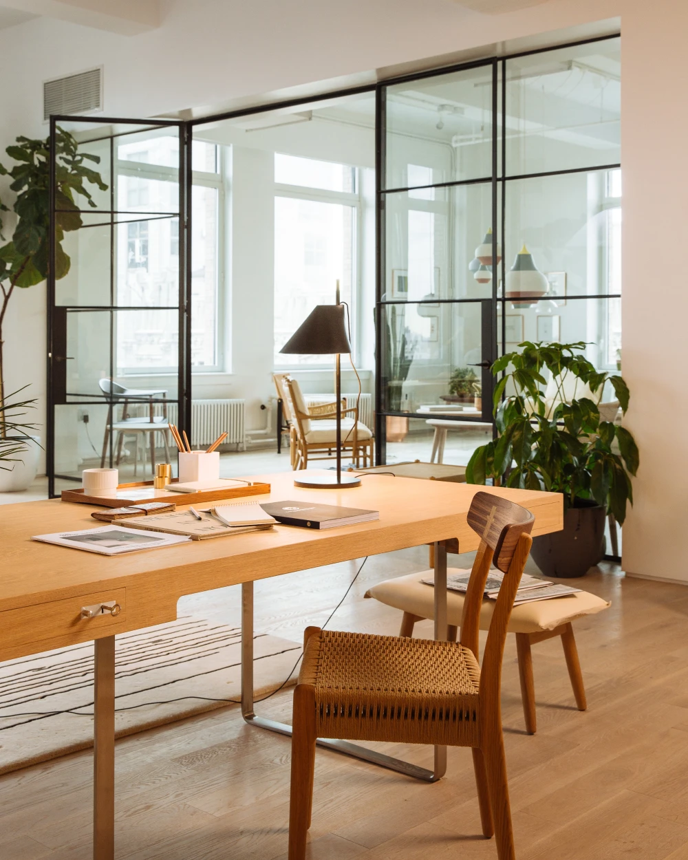 Een donkere AJ Floor Lamp staat naast het bureau; witte PH 5-hanglampen hangen in de lichte showroom met glazen wanden en houten meubels. © 2026 Louis Poulsen.