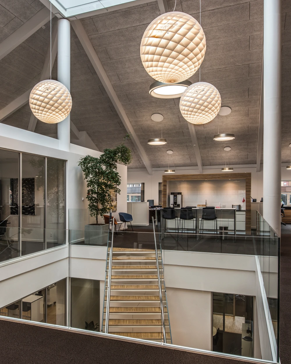 Witte Patera-hanglampen verlichten een modern kantooratrium met betonnen plafond, glazen balustrades en een trap. © 2026 Louis Poulsen.