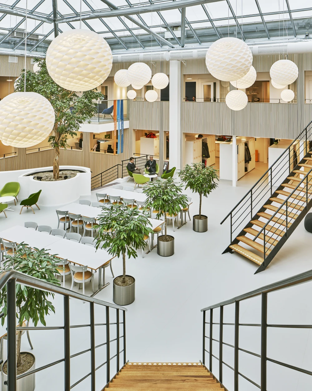 Witte Patera-hanglampen hangen op verschillende hoogtes boven zit- en eetgedeeltes in een licht kantooratrium. © 2026 Louis Poulsen.