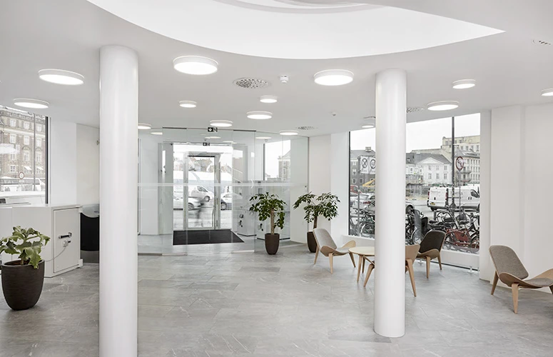 LP Circle Surface Mounted ceiling lights in Sydbank’s bright entrance, with a glass façade, grey tiled floor and seating. © 2026 Louis Poulsen.