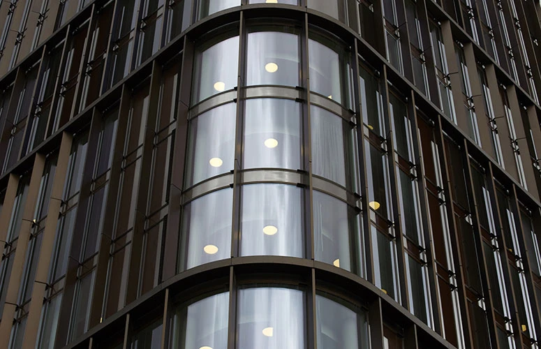 Round recessed ceiling lights glow behind the windows of a modern office building with a curved glass façade. © 2026 Louis Poulsen.