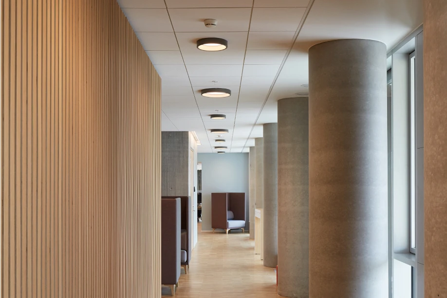 Ronde LP Circle Surface Mounted-plafondlampen verlichten een gang met een houten lattenwand, betonnen kolommen en ramen. © 2026 Louis Poulsen.
