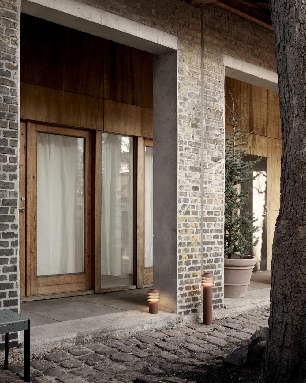 Bronzen Bysted Garden-bolderarmaturen verlichten een geplaveid pad bij een huis met een bakstenen gevel en houten deuren. © 2026 Louis Poulsen.