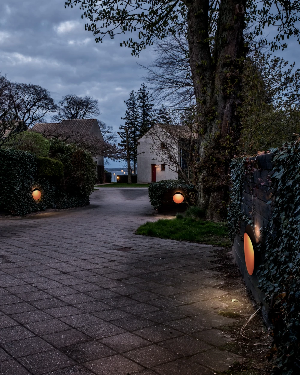 De Flindt-wandlamp geeft warm licht vanaf een tuinmuur naast een bestraat pad in de schemering. © 2026 Louis Poulsen.