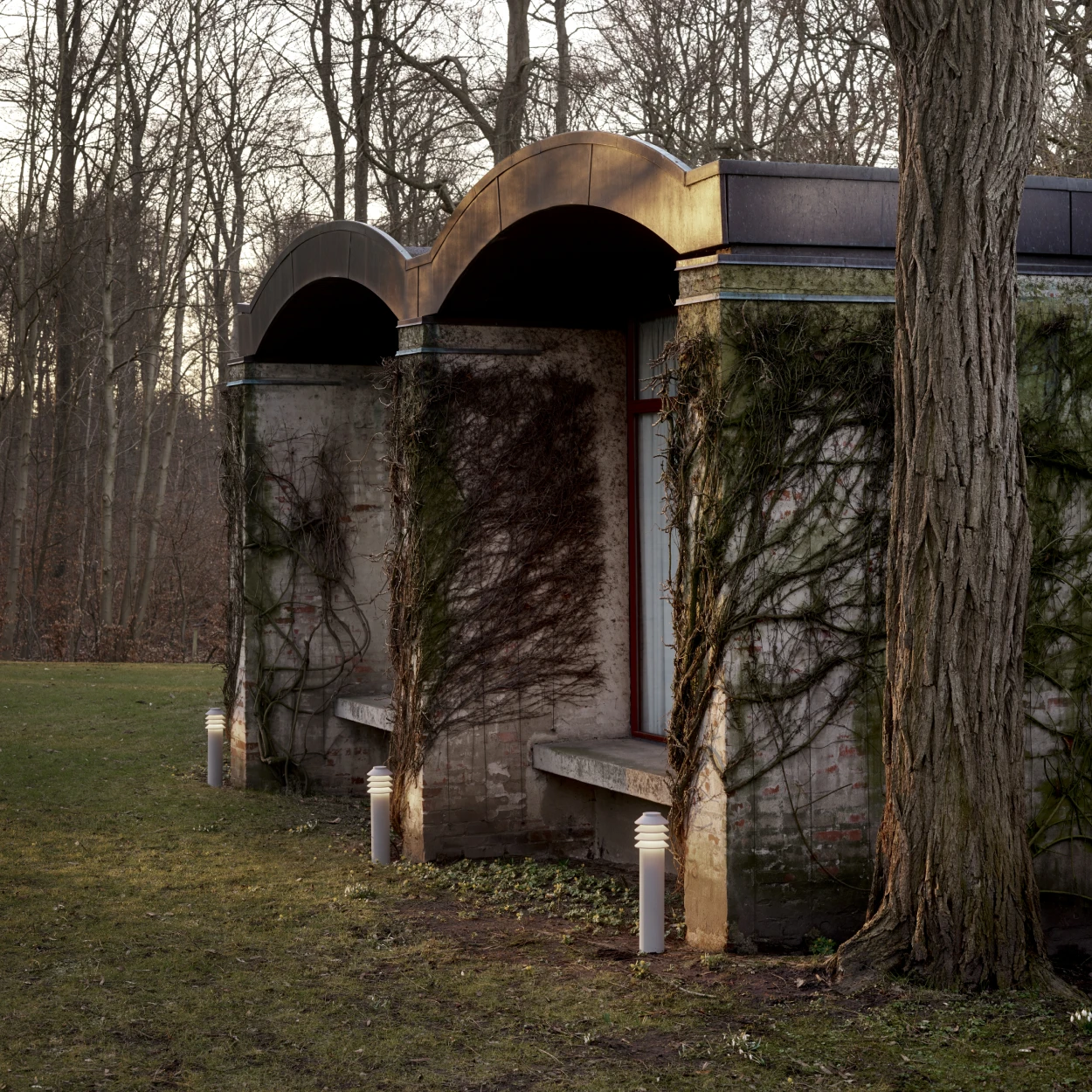 Albertslund Mini Post-bolderarmaturen verlichten een pad langs een met klimop begroeide muur in een bosrijke tuin. © 2026 Louis Poulsen.