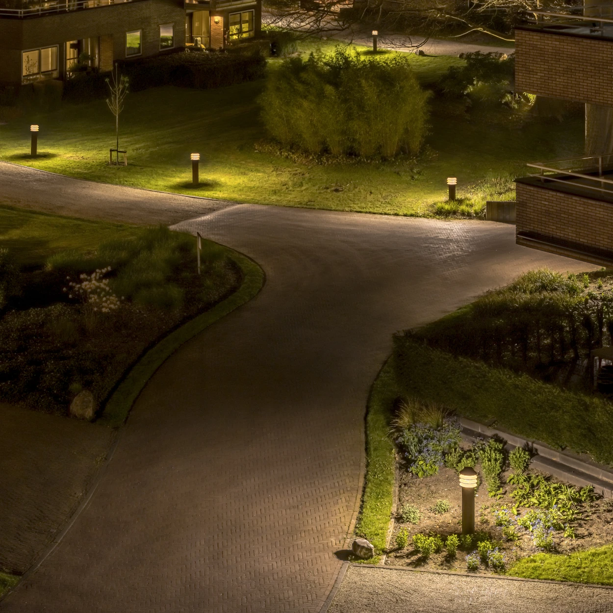 Albertslund Mini Post-bolderarmaturen verlichten 's avonds een gebogen pad en beplante perken bij woningen. © 2026 Louis Poulsen.