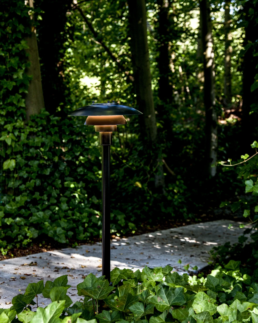 Een PH 3-2½-bolder met donkere afwerking verlicht een stenen pad in een weelderige tuin. © 2026 Louis Poulsen.