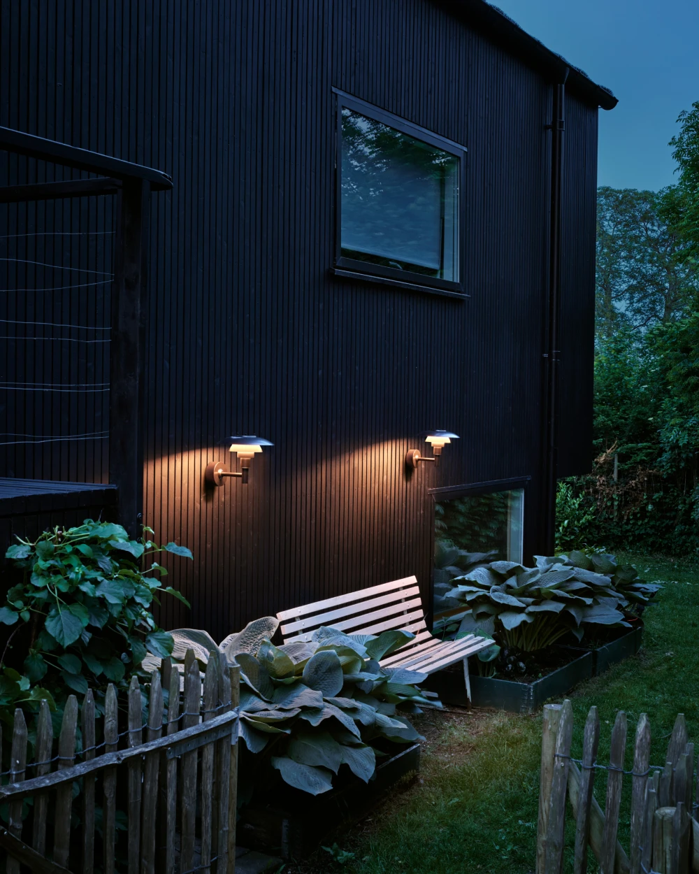 Twee PH 3-2½ buitenwandlampen werpen warm licht op de donkere houten gevel naast een tuinbank. © 2026 Louis Poulsen.