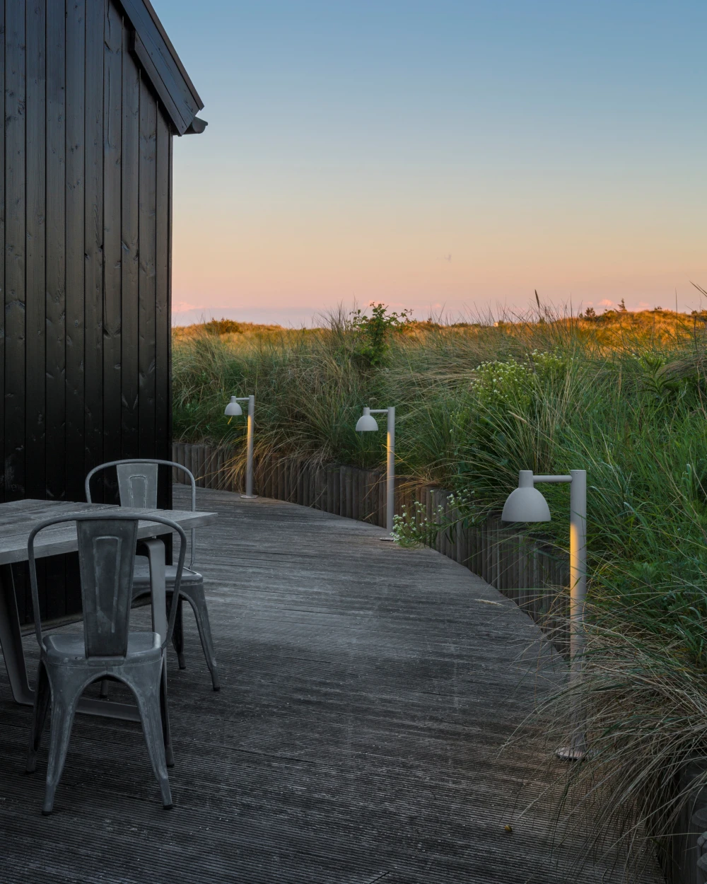 Toldbod 155-bolderarmaturen staan langs een houten terras bij een donker houten huis, tussen het helmgras in de avondschemering. © 2026 Louis Poulsen.