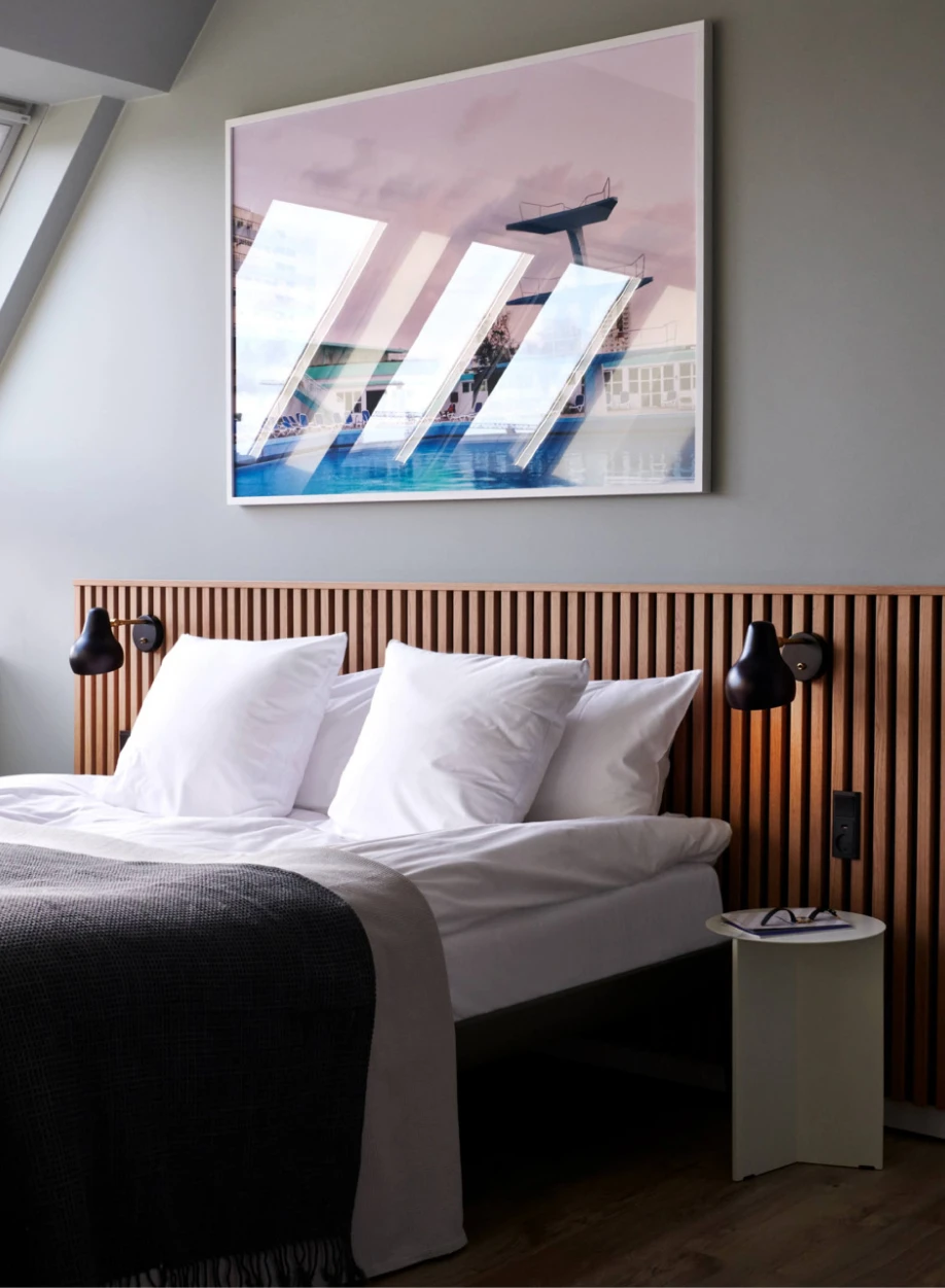 Black Keglen Wall Lamps flank a bed in a hotel room with a slatted wooden headboard. © 2026 Louis Poulsen.