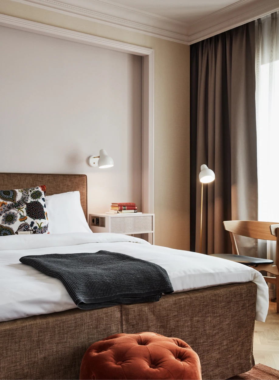 En vit AJ Wall Lamp och en vit AJ Floor Lamp lyser upp ett hotellrum med säng och mörka gardiner. © 2026 Louis Poulsen.